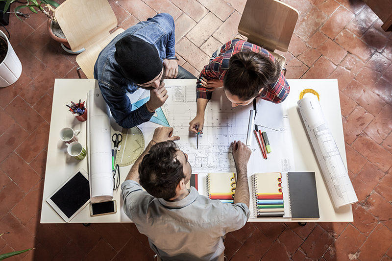 Two male and one female architects around a table working on blue prints for a building design Two male and one female architects around a table working on blue prints for a building design